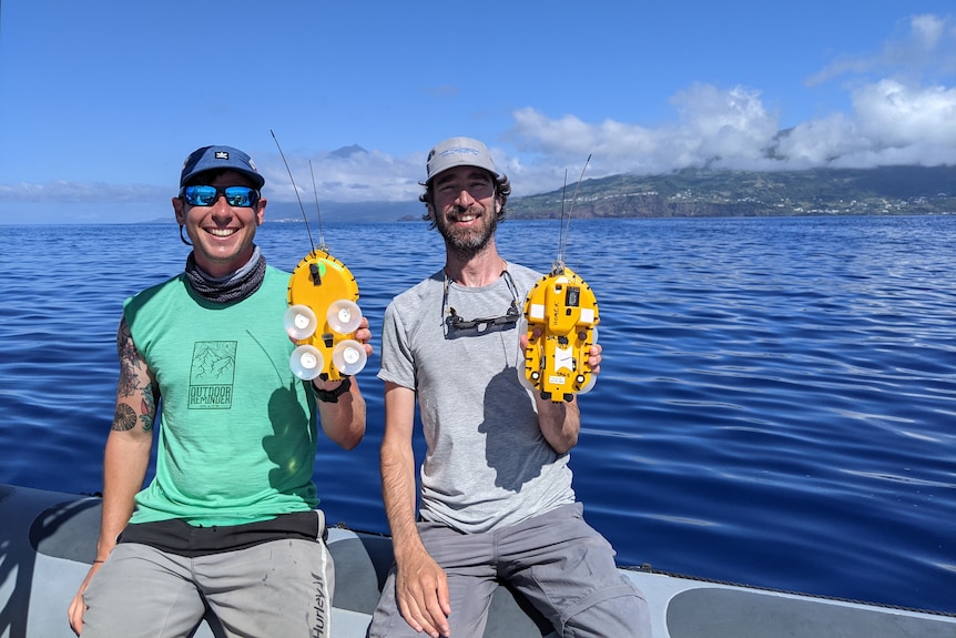 Two men with caps and t-shirts sitting on a boat near an island with large yellow oval objects held in their left hands.
