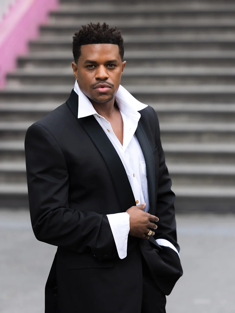 Person in a black suit with a white, open-collar shirt stands confidently in front of steps, posing
