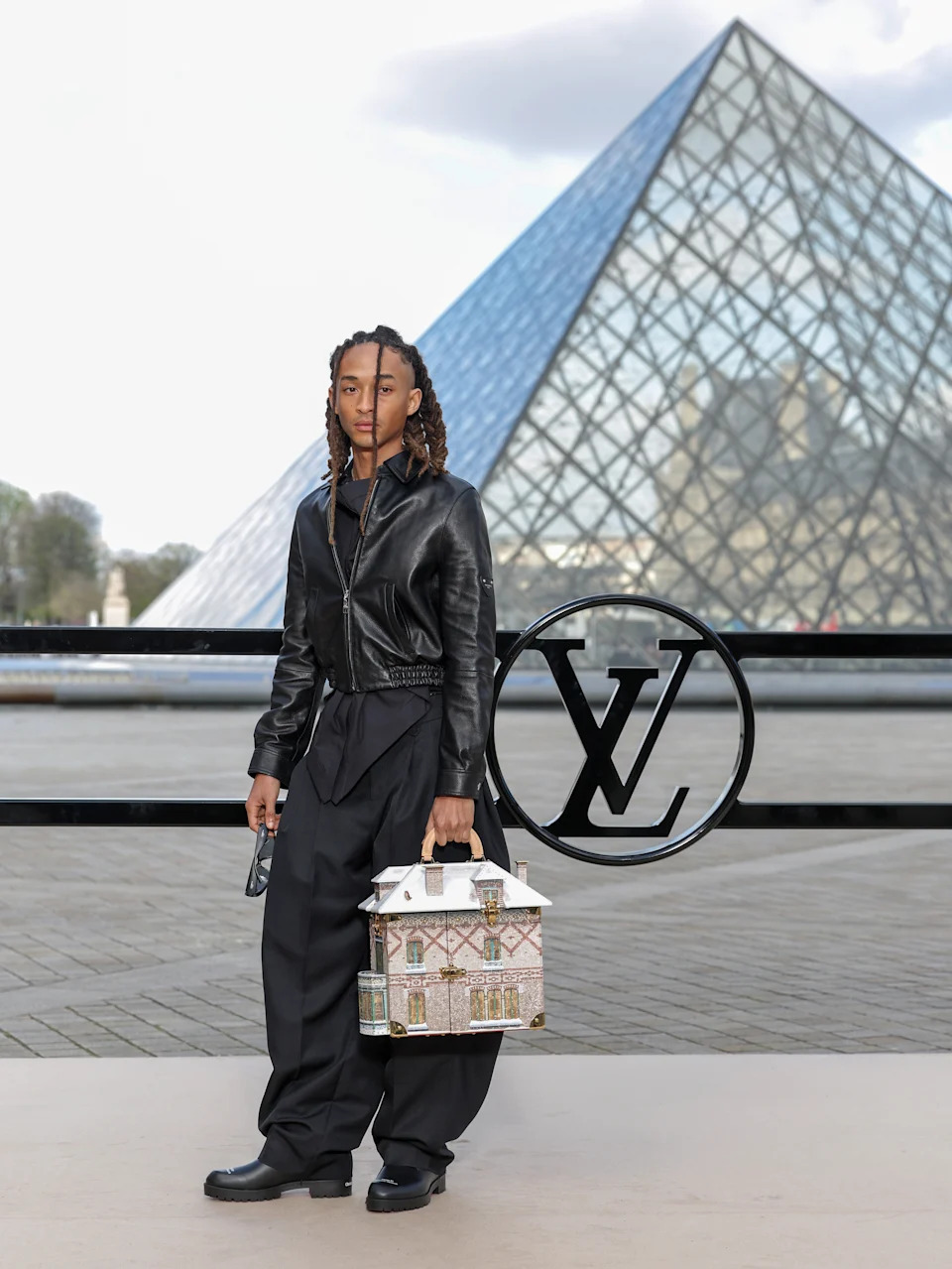 Person in leather jacket and wide-leg pants poses near a pyramid glass structure, holding a handbag shaped like a small house