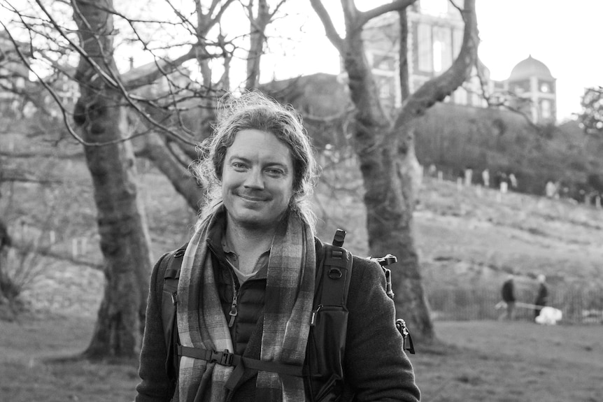 A black-and-white photo of a man with his hair tied back, wearing a scarf, warm jacked and a backpack, standing in a field.