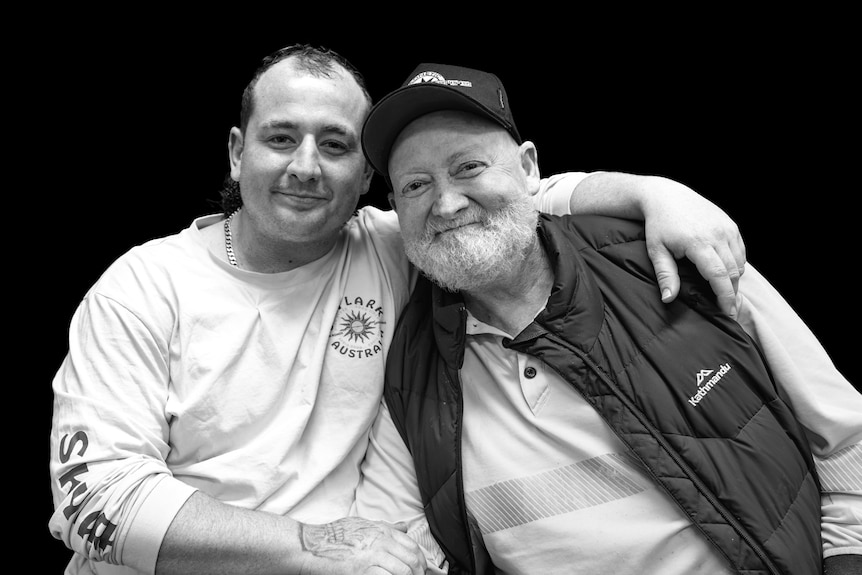 A man has his arm around an older man with a white beard, as both men smile at the camera.