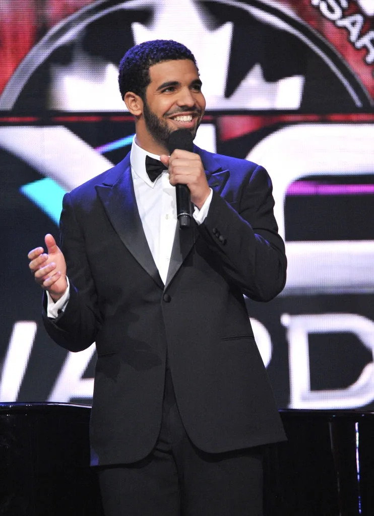 A man in a black tuxedo with a bow tie holds a microphone, smiling on stage at an awards event
