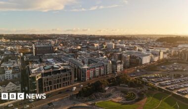 A drone shot showing St Helier from the sky. The sun is low and it gives a golden glow to the buildings packed together. There is a park in the foreground and a car park next to it.
