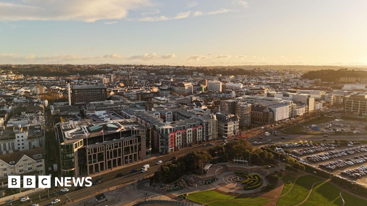 A drone shot showing St Helier from the sky. The sun is low and it gives a golden glow to the buildings packed together. There is a park in the foreground and a car park next to it.