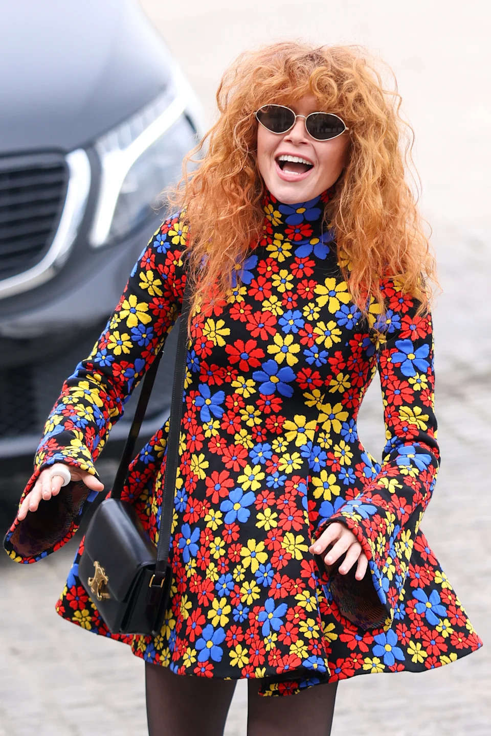 Person in a vibrant floral-patterned dress with flared sleeves, wearing cat-eye sunglasses, smiling