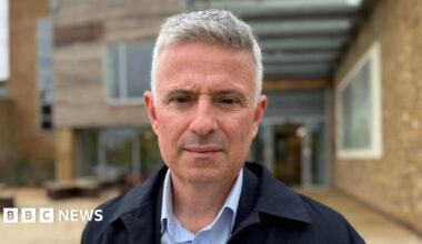 Paul Scott has short grey hair and is shaven. He wears a light blue shirt and navy blue jacket, and is standing outside a modern building.