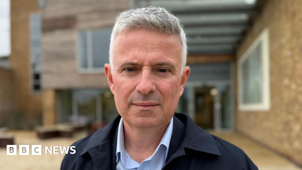 Paul Scott has short grey hair and is shaven. He wears a light blue shirt and navy blue jacket, and is standing outside a modern building.