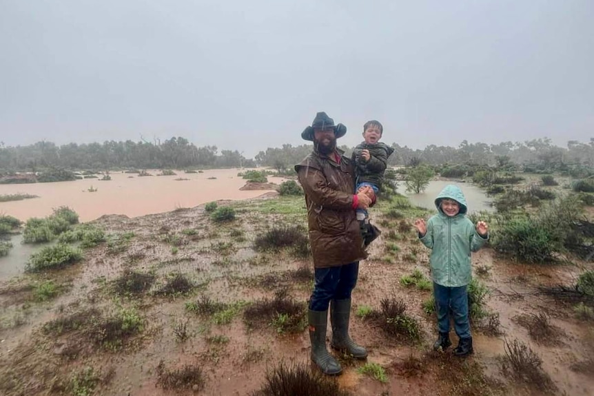 A grazier in an akubra stands in a flooded paddock with his two young children