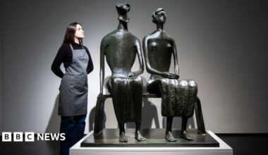 A bronze sculpture of a couple sitting on a bench. A woman in a black top and grey apron stands next to the sculpture with her hands behind her back, looking at it.