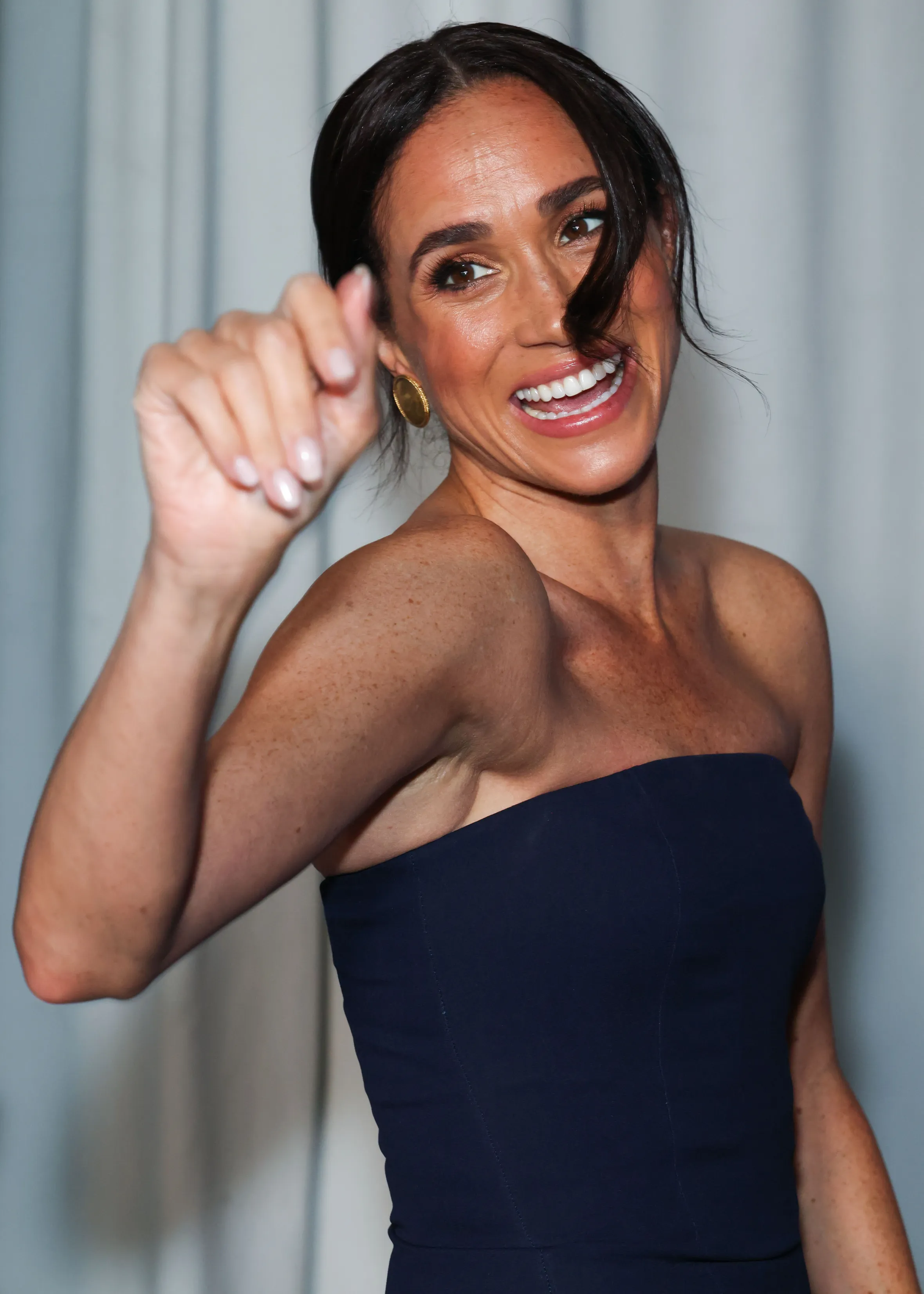 Meghan, Duchess of Sussex, laughing and gesturing, wearing a dark blue strapless dress.