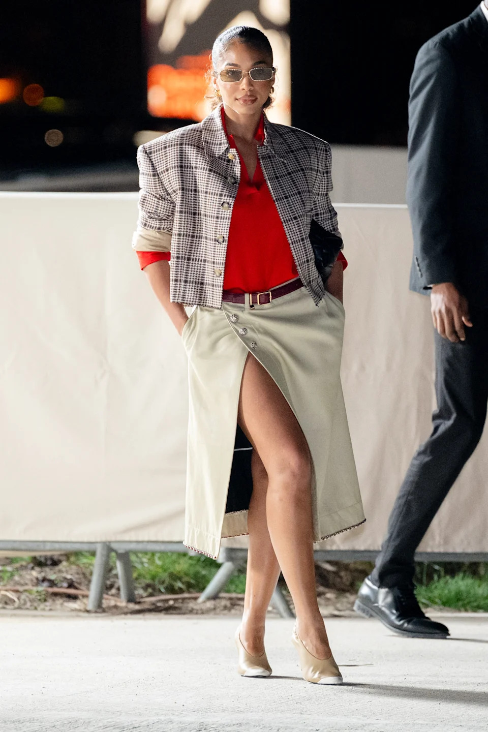 Celebrity in a plaid blazer, red blouse, and beige knee-length skirt with a high slit, walking outdoors at an event