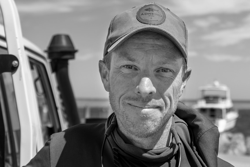 A black-and-white photo of a man wearing a baseball cap and a hoody shirt, standing next to his car.