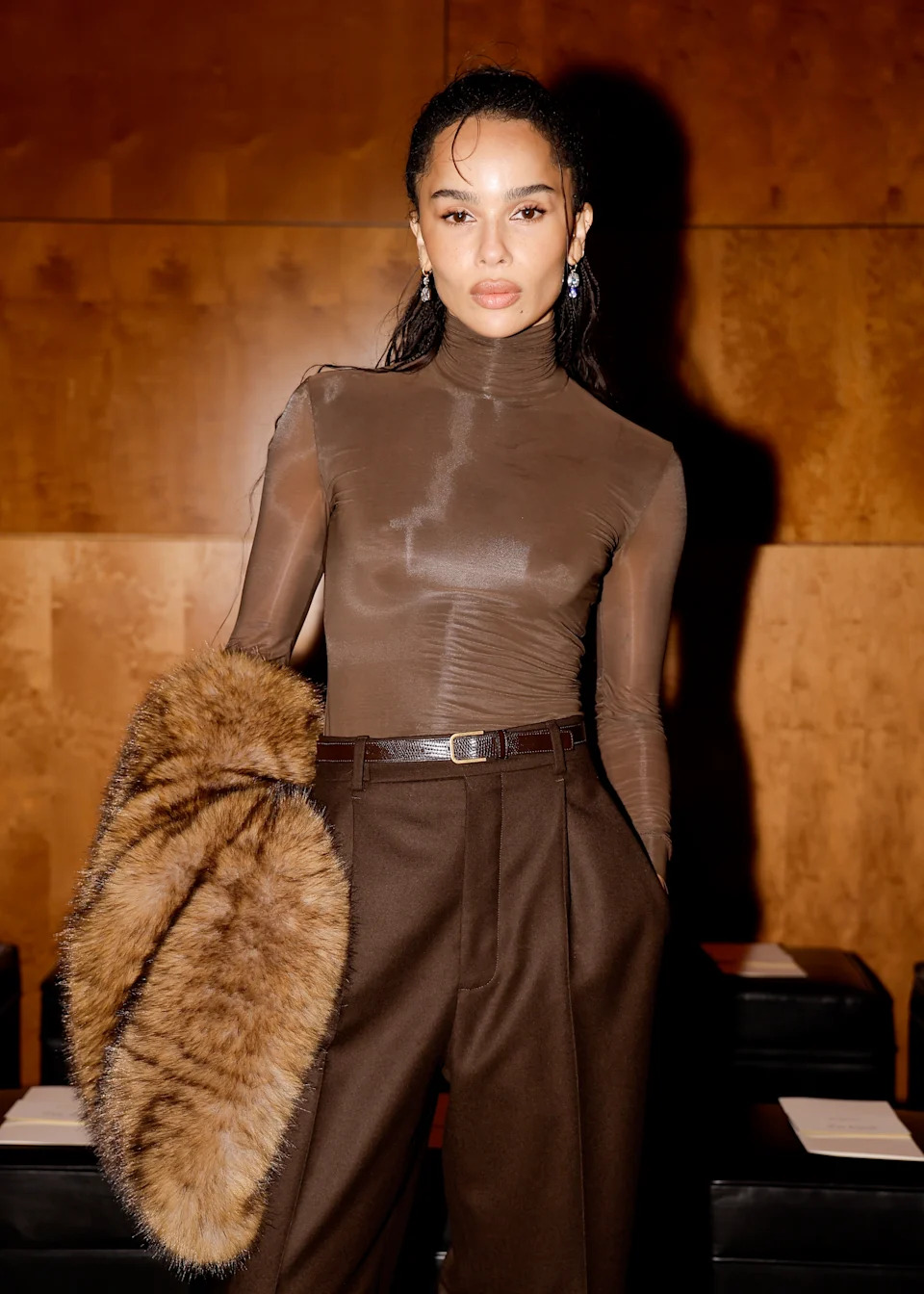 A person poses confidently in a sheer turtleneck top and high-waisted trousers, holding a faux fur accessory at a celebrity event