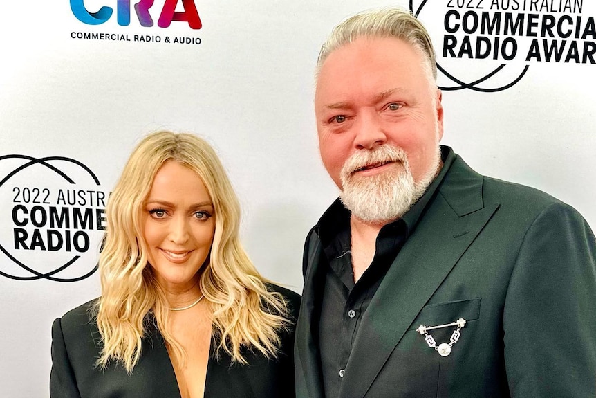 A blonde woman and man with a grey beard wearing black suits stand in front of a media wall.