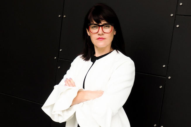 A woman with straight black hair, glasses and a white jacket has her arms folded and looks straight ahead.