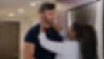 A woman embraces a smiling man in a hallway, both appear happy. A modern black-and-white painting decorates the wall behind them.