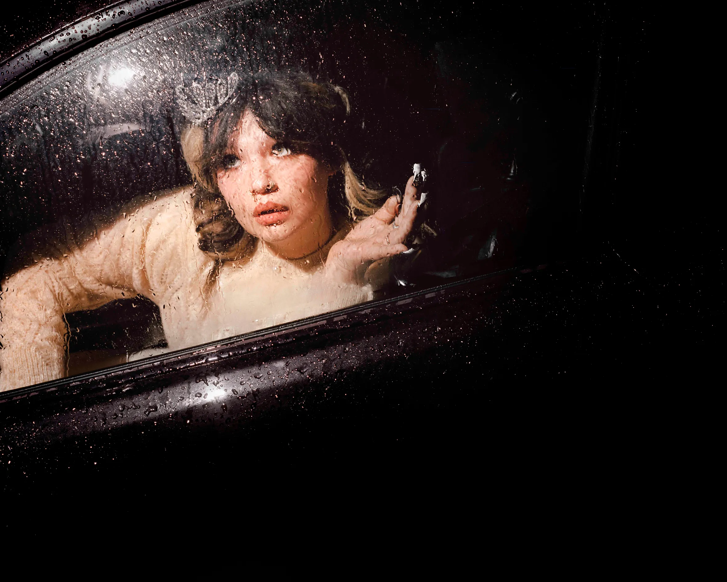 Lola Young looks out a car window covered in raindrops at night.