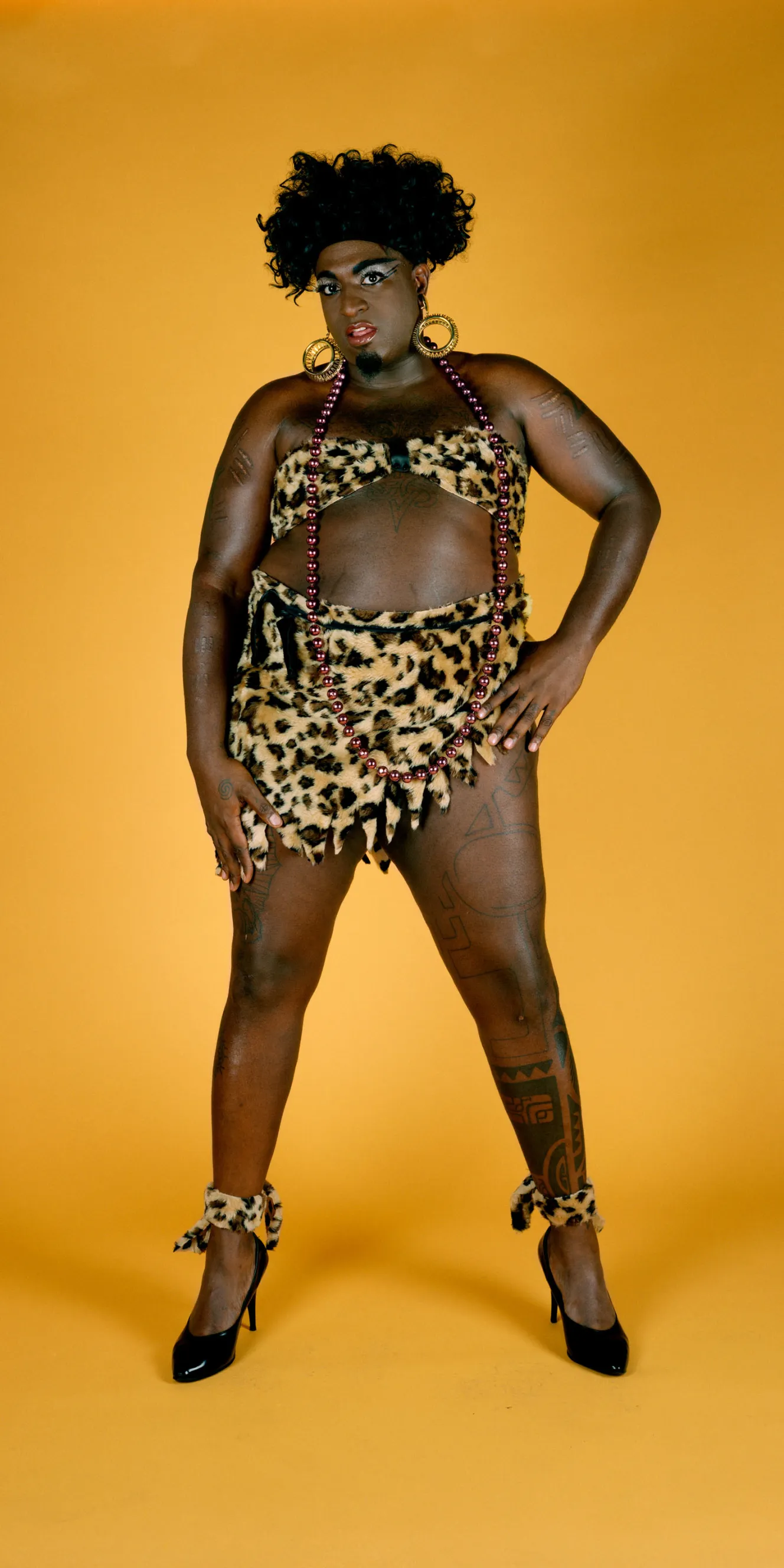 A person in drag, Divinity Fudge, poses in a leopard print bikini, long red beaded necklace, large gold hoop earrings, black high heels, and leopard print ankle cuffs.