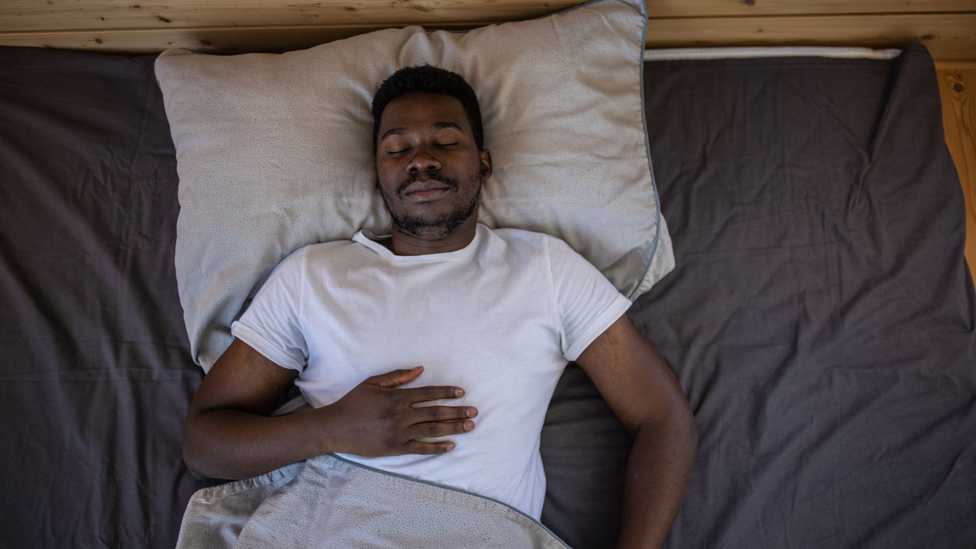 A man lies on his back in bed with his eyes closed and one hand resting on his chest.