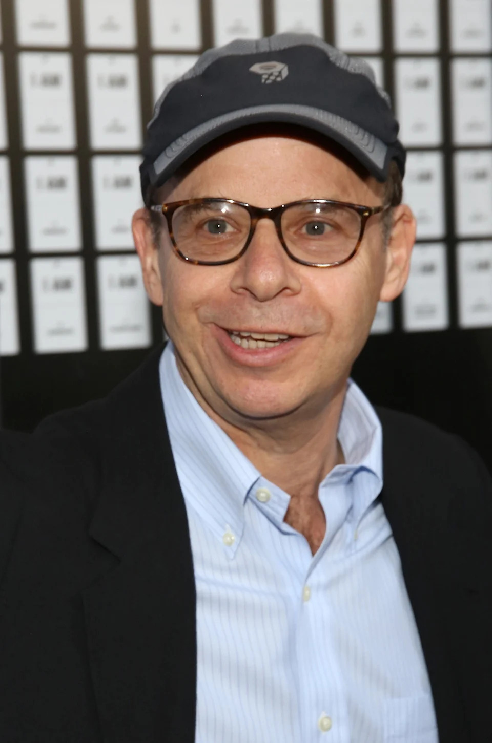 Rick Moranis in glasses and a cap smiling at an event, wearing a blazer over a button-up shirt