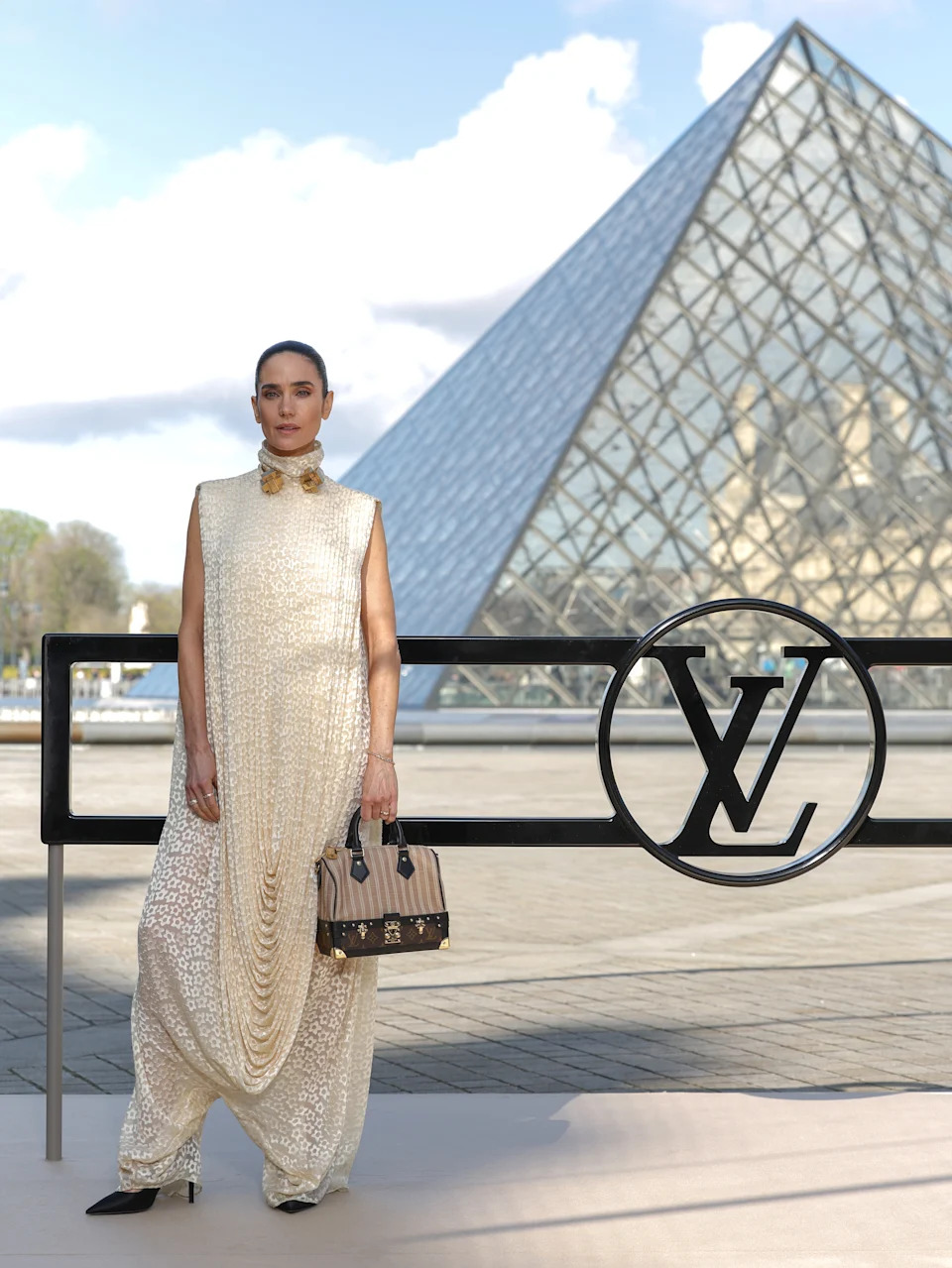 Celebrity in a flowing, textured gown poses near a glass pyramid structure, holding a designer bag at a fashion event