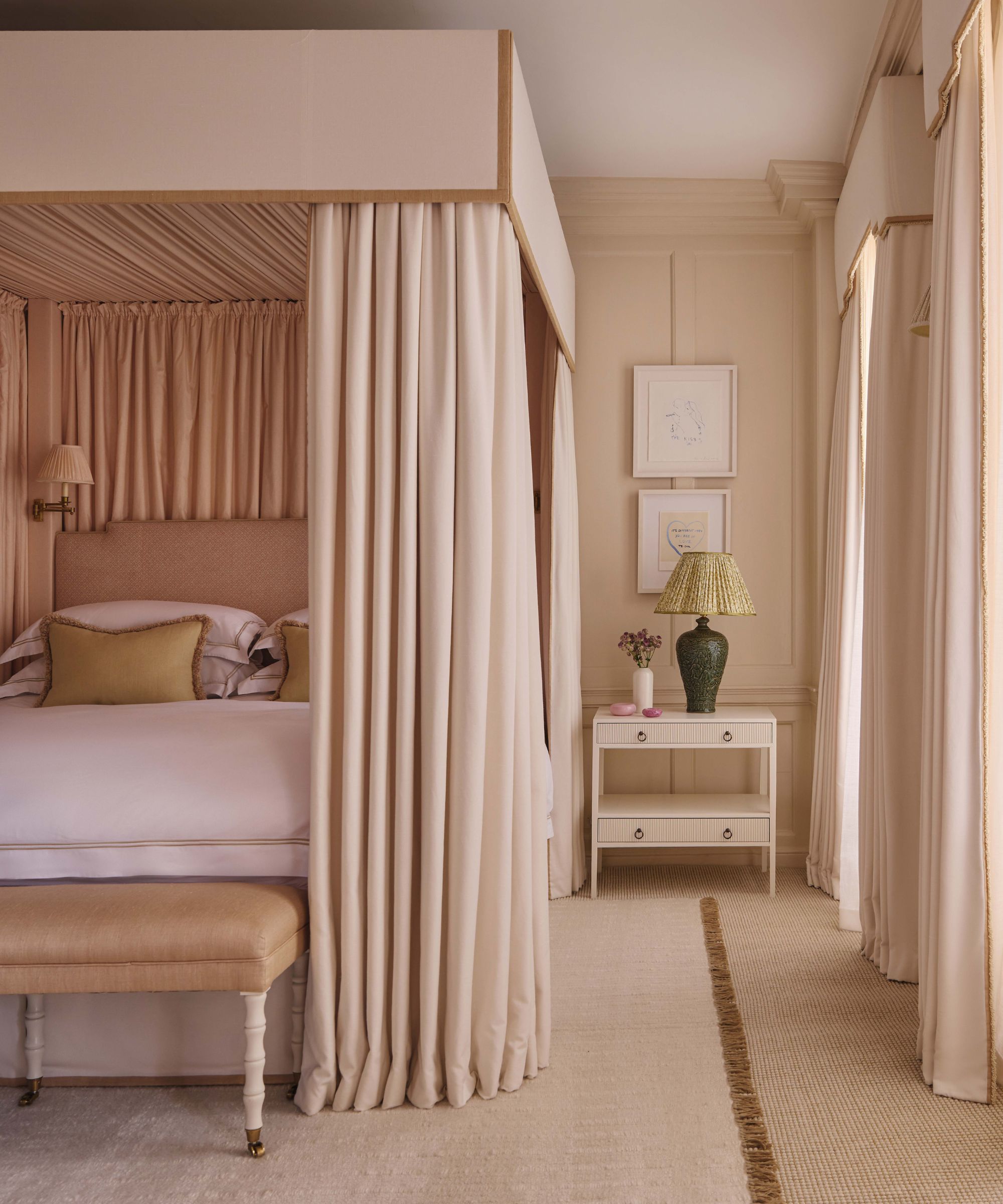 A serene, monochromatic bedroom featuring a grand four-poster bed draped in heavy cream fabric. The room is styled with a matching cream nightstand, a green ceramic lamp with a pleated shade, and soft neutral wall paneling.