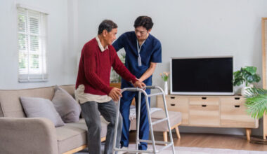 Caring nurse smiling while assisting elderly man with walker in sunny living room