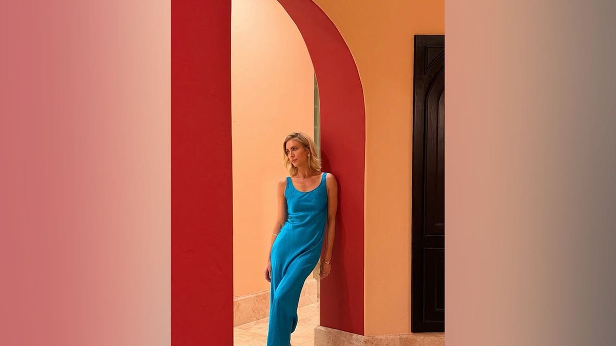 Brittany Snow standing in a doorway wearing a bright blue silk slip dress with a high leg slit.