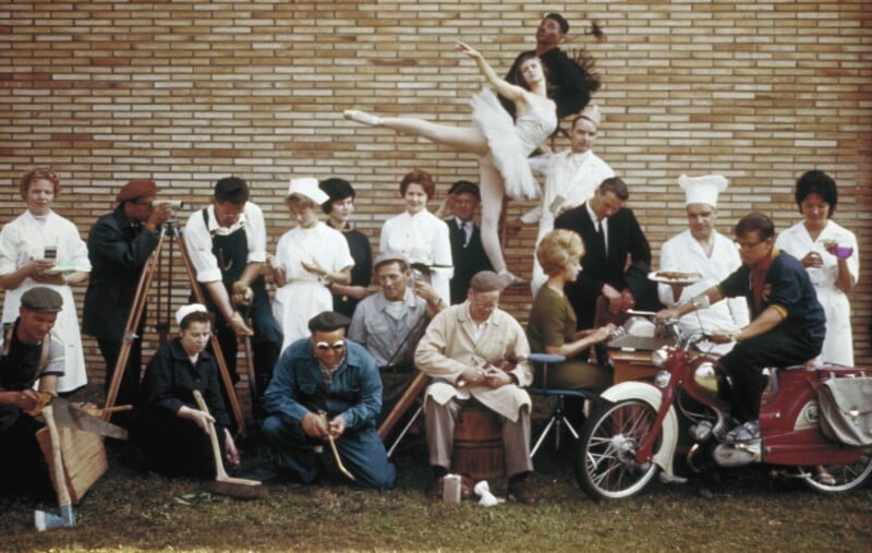 A group of people in work uniforms, including chefs, nurses, and laborers, pose in front of a brick wall. A ballerina stands on pointe above them, and a man sits on a red motorbike in the foreground.