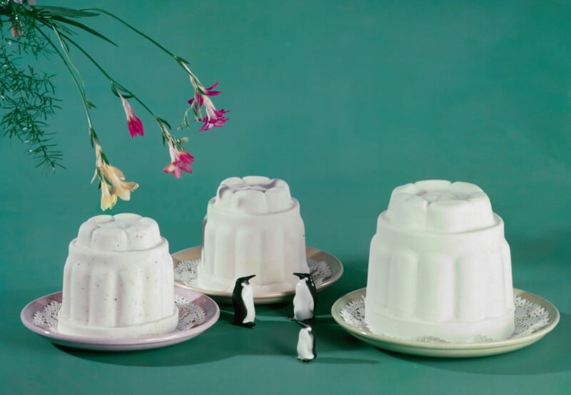 Three white molded desserts on plates are displayed against a teal background, with three miniature penguin figurines standing in front. Pink and yellow flowers hang from the top left corner.