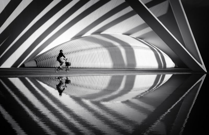 A person rides a bicycle past a modern, curved building with dramatic striped shadows, reflected perfectly in a pool of water below. The image is in black and white.