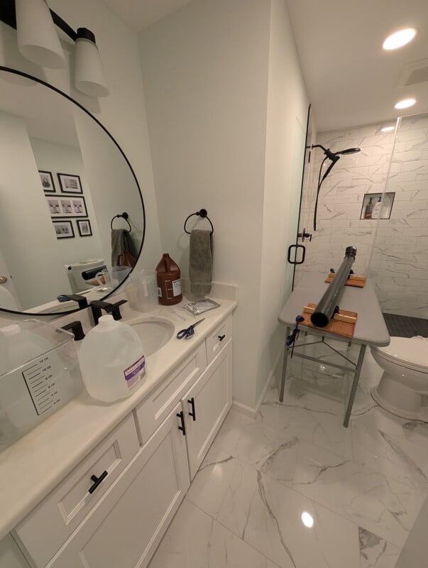 A bathroom with white cabinets and marble floor, a round mirror above the sink, cleaning supplies on the counter, and a folding table set up over the toilet. The shower area has a glass door and white tiled walls.