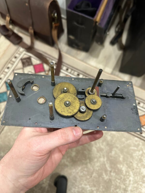 A hand holds a metal plate with several interlocking brass gears and rods, possibly part of a clock or mechanical device, against a background with a patterned rug and leather bags.