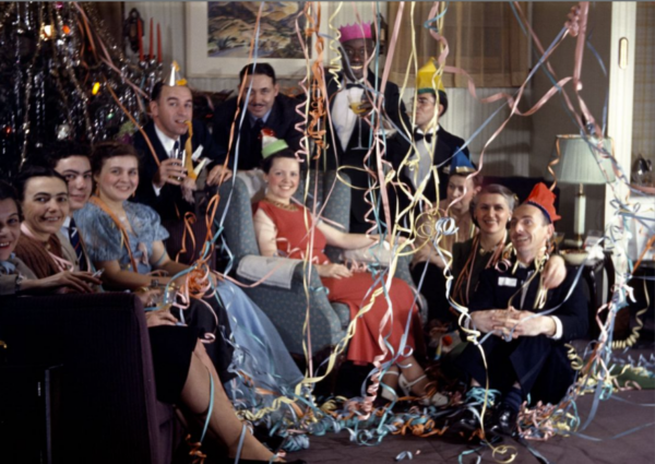 A group of people in festive hats celebrate indoors, smiling and sitting close together, surrounded by colorful streamers and decorations, creating a lively party atmosphere.