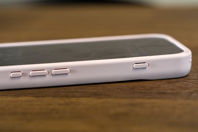 A close-up side view of a smartphone with a white or light pink case, showing the volume and power buttons, resting on a wooden surface.