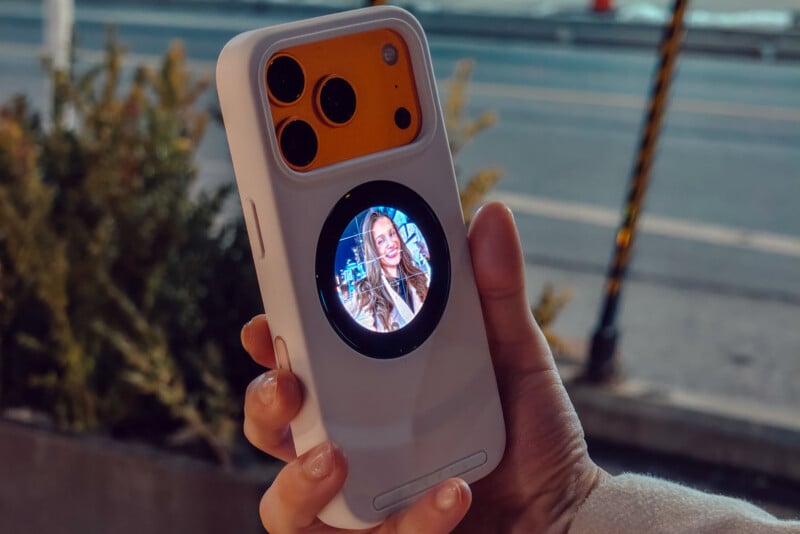A hand holds a smartphone with a unique white case featuring a small circular screen on the back, displaying a smiling woman, while the main camera lenses appear above the screen. The background shows a street and some greenery.