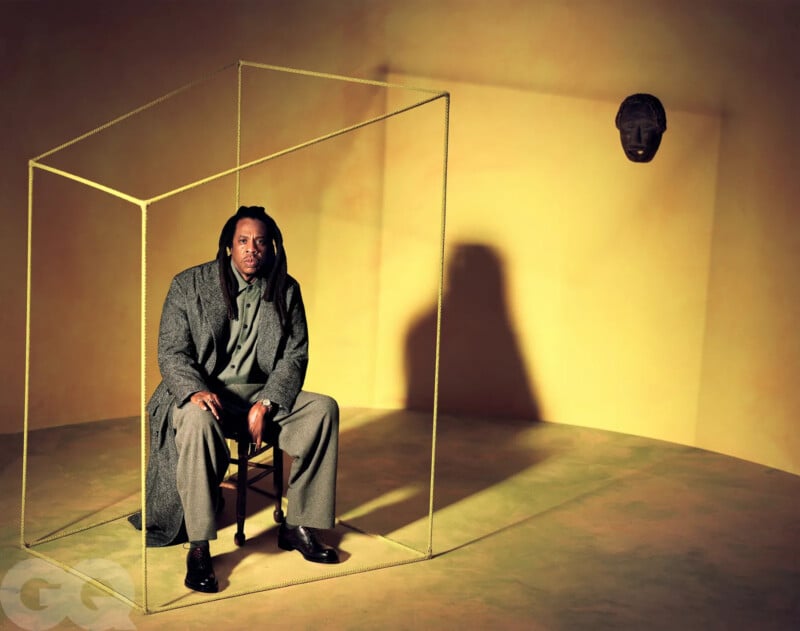 A man in a gray suit with long hair sits on a wooden chair inside a minimal yellow wireframe cube, set against a yellow background. A carved mask hangs on the wall to his right, casting shadows.