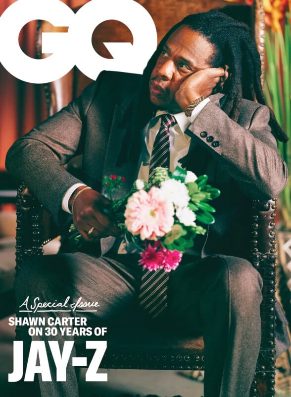 Jay-Z, dressed in a brown suit, sits on a chair holding a bouquet of flowers. He rests his head on his hand, looking thoughtful. The GQ logo and text about a special issue on 30 years of Jay-Z are visible.