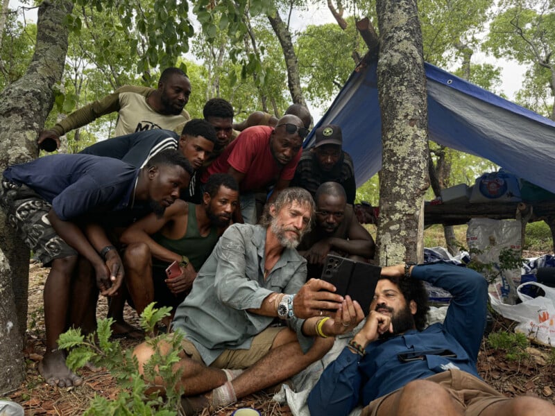 A group of men gather closely under a makeshift tarp in a forest, intently looking at a smartphone held by two men in the foreground, who appear relaxed and engaged. Supplies and bags are scattered around them.