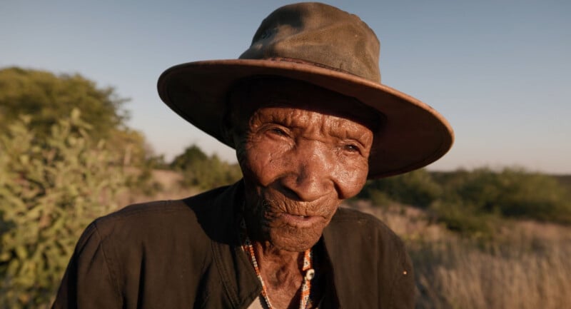 An elderly person with deeply wrinkled skin, wearing a wide-brimmed hat and a dark jacket, stands outdoors in the sunlight with dry grass and greenery in the background.