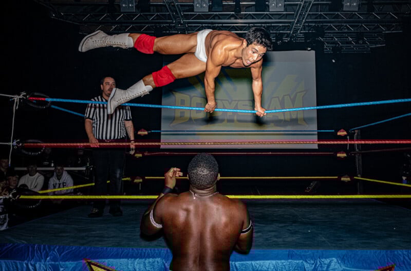A wrestler in white trunks and red knee pads leaps mid-air off the ropes toward another wrestler standing in the ring as a referee and audience watch under dramatic lighting.