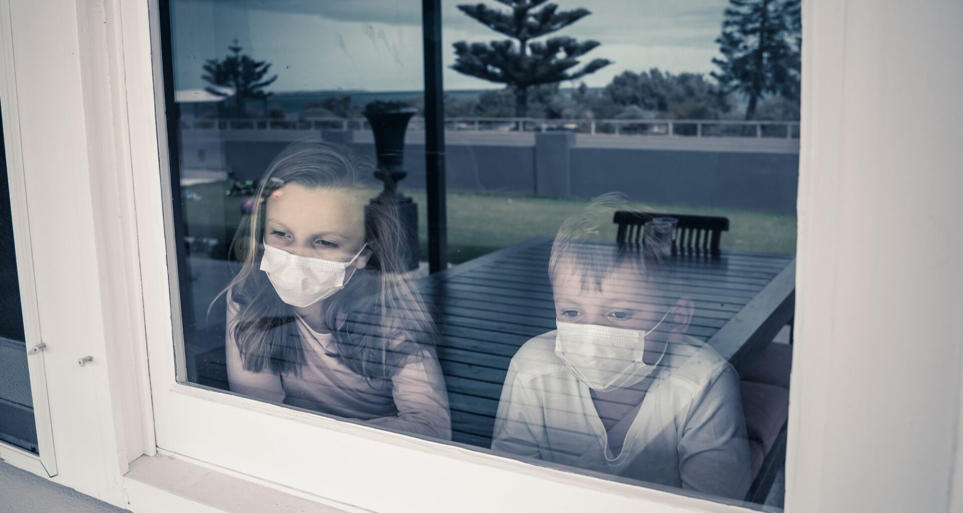 COVID-19 Lockdowns. Sad children with face mask looking through the window during quarantine at home. Kids feeling sad as school is closed. Coronavirus outbreak isolation and kids mental health.