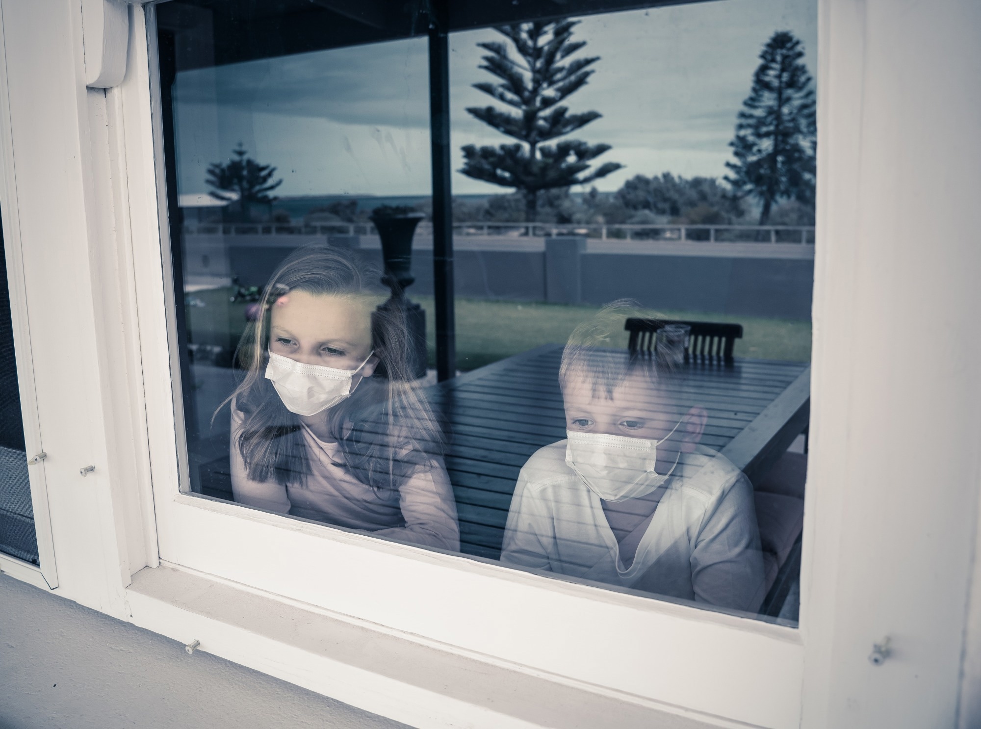 COVID-19 Lockdowns. Sad children with face mask looking through the window during quarantine at home. Kids feeling sad as school is closed. Coronavirus outbreak isolation and kids mental health.