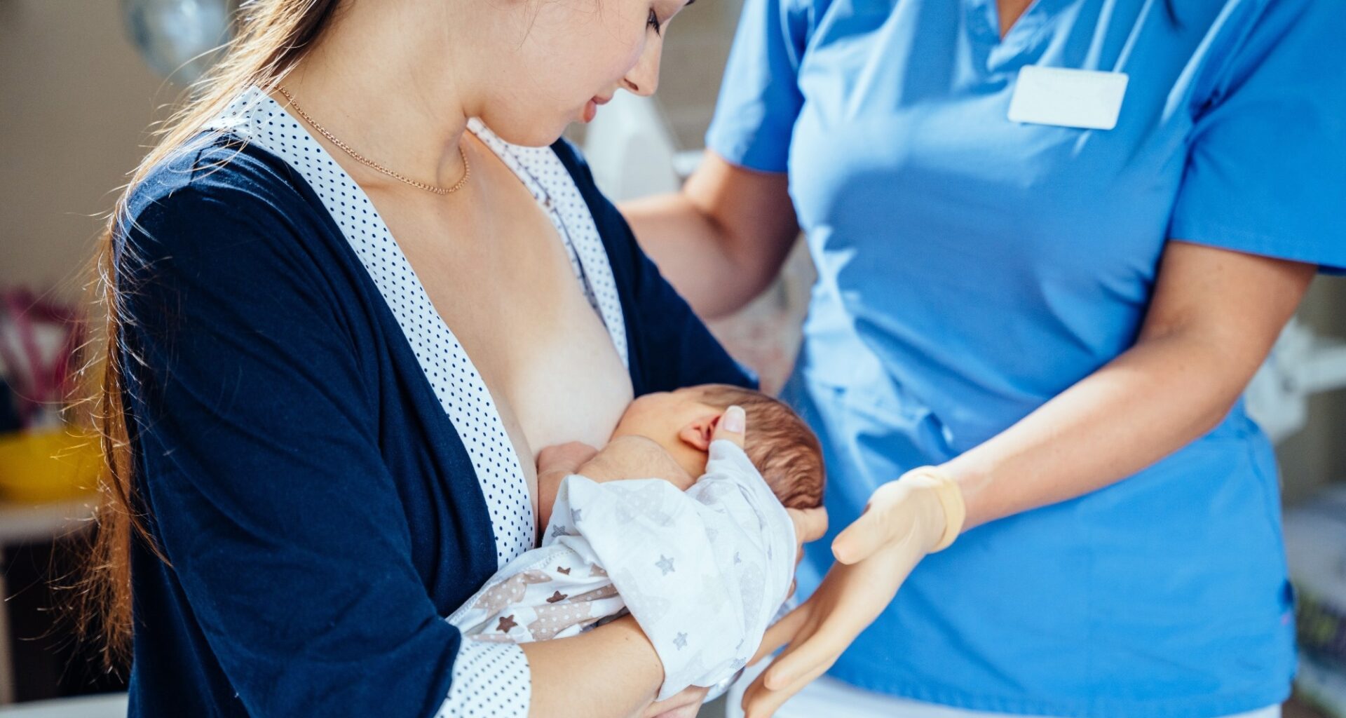 Unrecognizable midwife or breastfeeding consultant supporting a breastfeeding mother with her newborn baby girl in hospital ward.