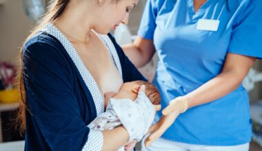 Unrecognizable midwife or breastfeeding consultant supporting a breastfeeding mother with her newborn baby girl in hospital ward.