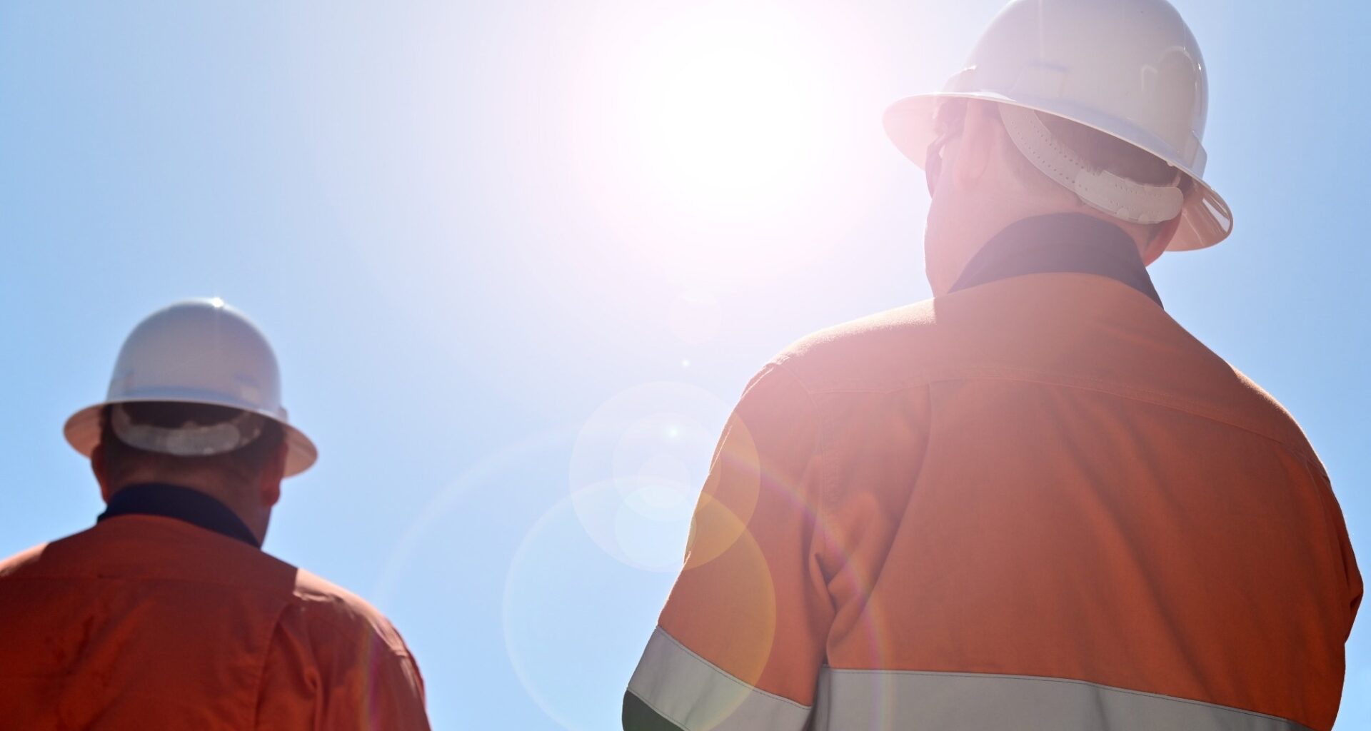 Two unrecognizable blue collar workers isolated against clear blue sky with sun flare.