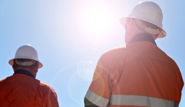 Two unrecognizable blue collar workers isolated against clear blue sky with sun flare.