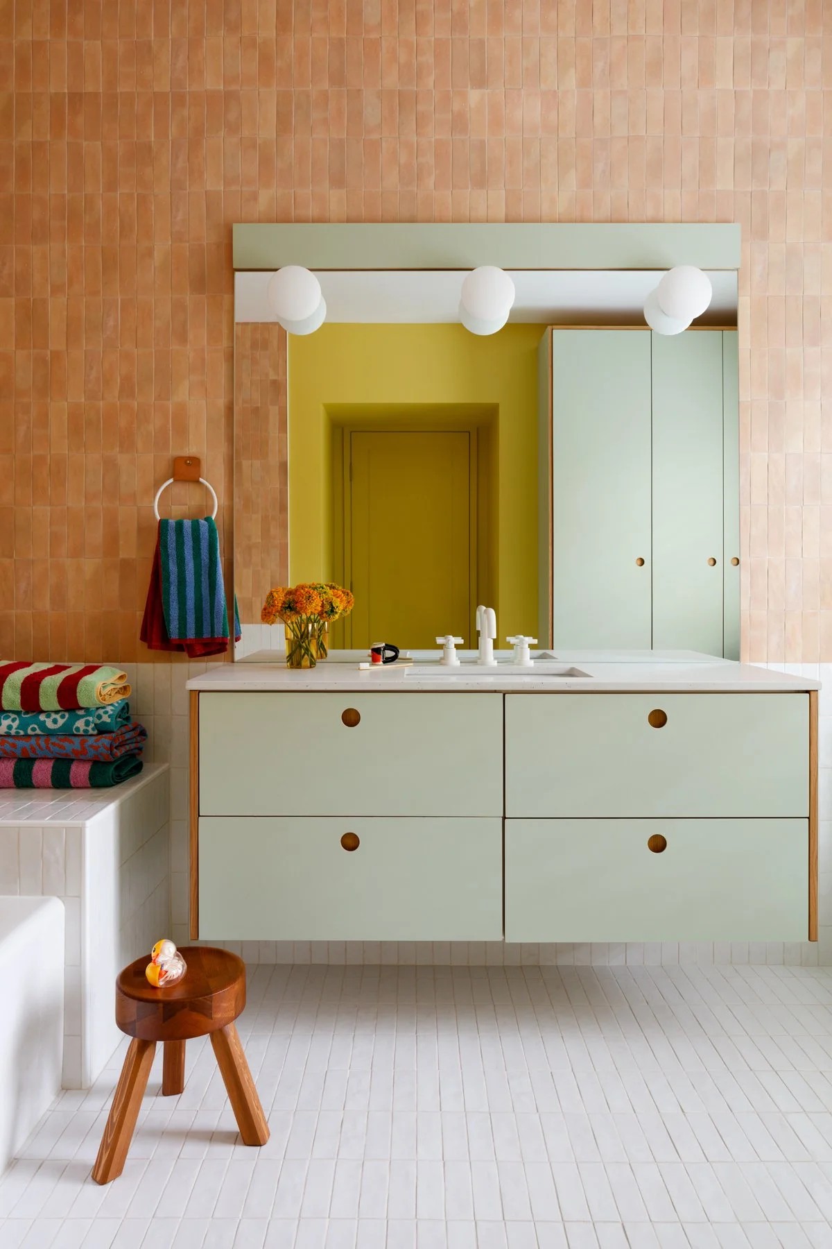 A bathroom with peach tiles on the wall and white tiles on the ground. There is a large floating sink and counter unit with mint green cabinets. A large mirror hangs above the sink with bulb sconce lights.