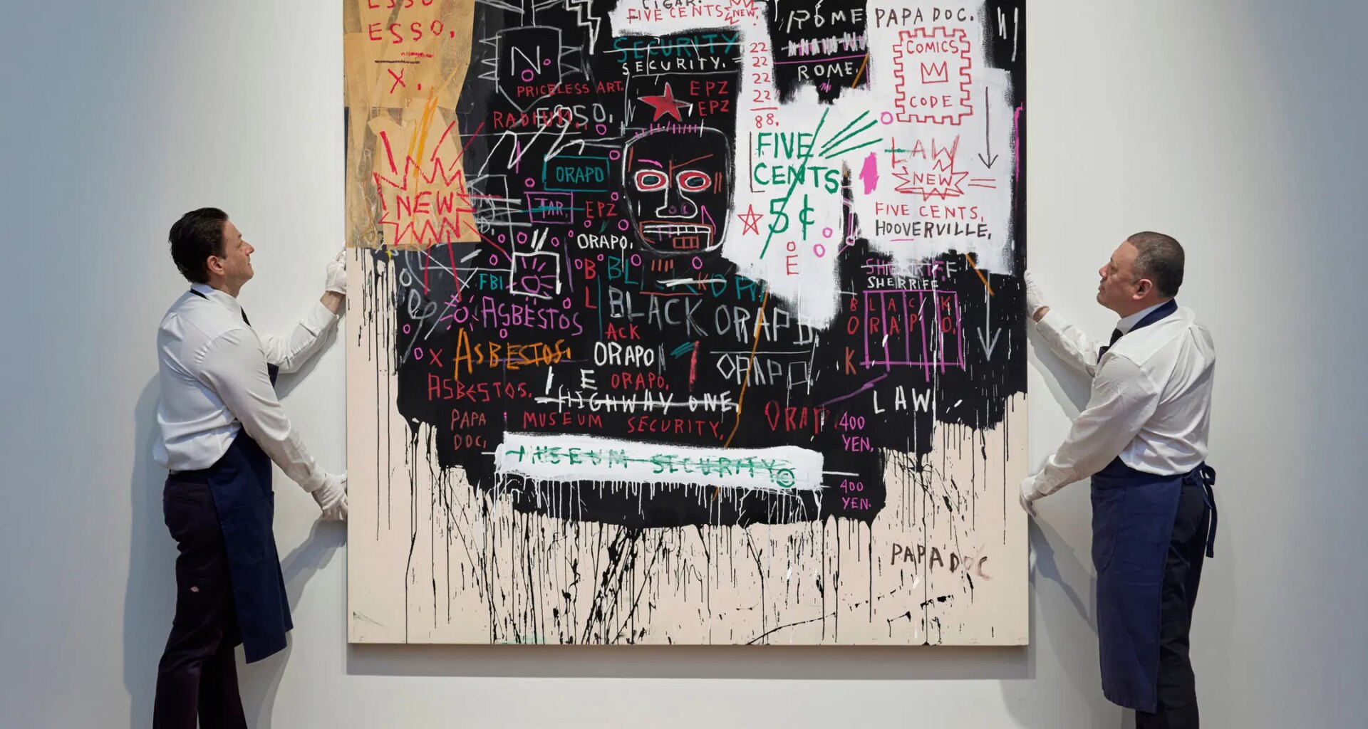 Two men in aprons adjusting a large abstract painting with various words, numbers, and symbols on a white gallery wall.