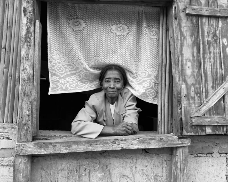 An elderly woman with short hair leans on the ledge of an open wooden window, framed by a lace curtain. She wears a light jacket and smiles gently, looking directly at the camera. The photo is in black and white.